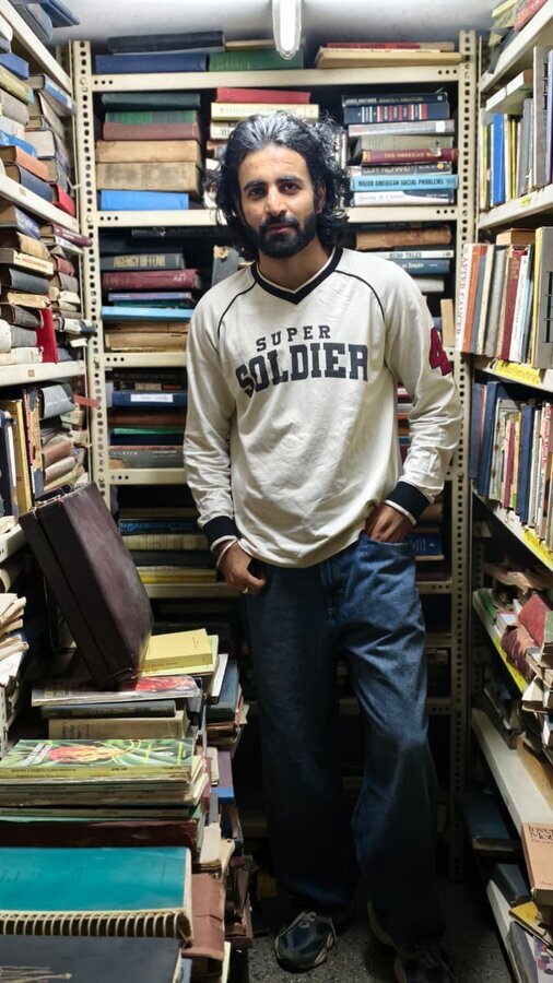 Aman standing in a cramped secondhand bookstore aisle, stacks of old books on every shelf